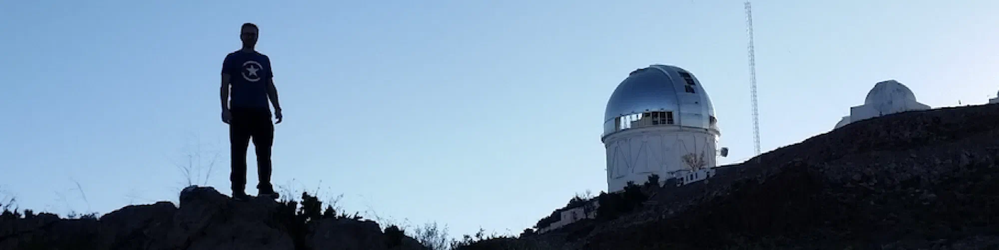 John Moustakas silhouetted on a ridge beside a telescope dome at dusk